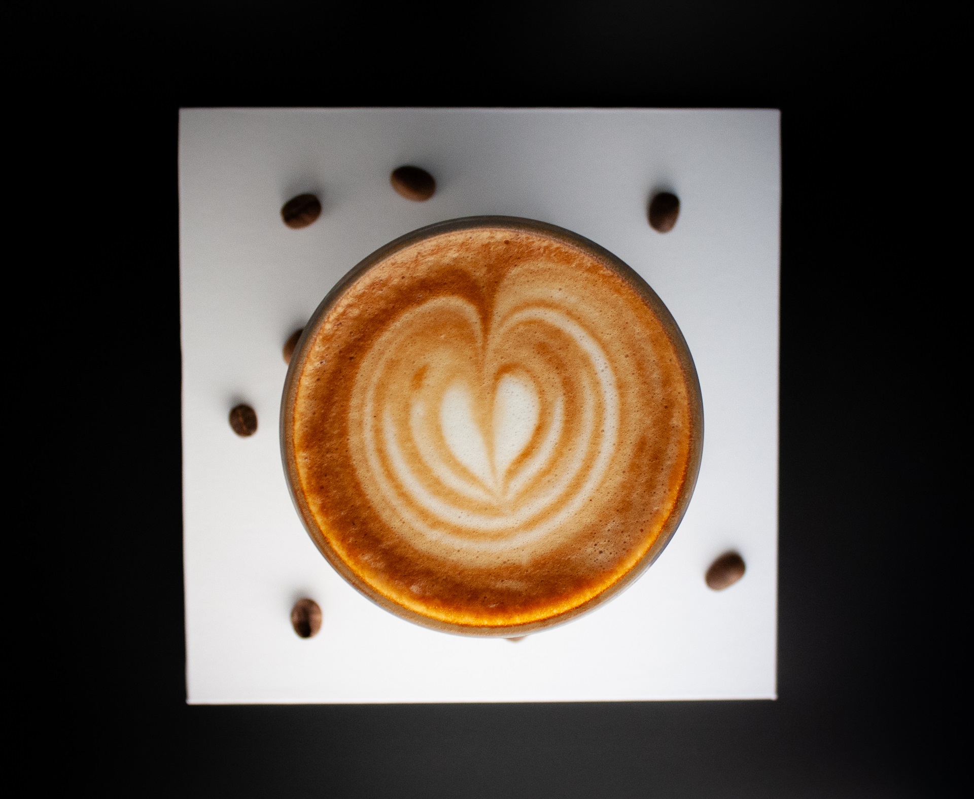 Birds Eye view of a cup off coffee on a white square surrounded by coffee beans